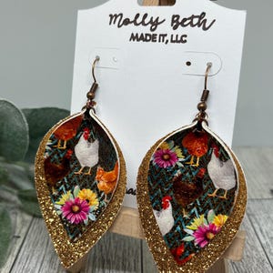 Könnte beinhalten: Goldene Glitzer-Tropfenohrringe mit einem mehrschichtigen Design mit einem floralen und Hühnerdruck. Die Ohrringe werden von Molly Beth Made It, LLC hergestellt.