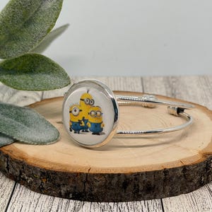 Puede incluir: Un brazalete de tono plateado que presenta un dije circular con fondo blanco y un estampado gráfico de tres personajes Minion amarillos con overoles azules y gafas. El brazalete tiene una banda de metal pulido delgada y un engaste redondo.