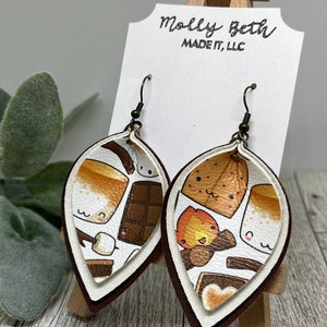 May include: A pair of brown leather earrings with a white layer featuring a s'mores design. The earrings are shaped like a teardrop and have a gold hook.