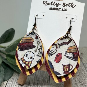 May include: A pair of dangle earrings with a Harry Potter theme. The earrings are made of faux leather and feature a layered design with a white background and red and gold stripes. The top layer features a variety of Harry Potter themed images, including a book, a key, a cup, a red gem, and a white owl with a scarf. The earrings are labeled "Molly Beth Made It, LLC."