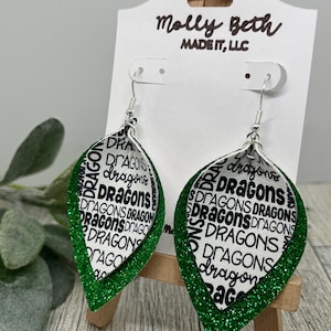 May include: A pair of leaf-shaped earrings with a white background and the word "Dragons" in black. The earrings have a green glitter border and silver hooks. The earrings are displayed on a white card with the text "Molly Beth MADE IT, LLC".