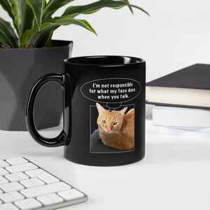 Könnte beinhalten: Schwarze Kaffeetasse mit einer weißen Grafik einer Katze mit dem Text "Ich bin nicht verantwortlich für das, was mein Gesicht tut, wenn du sprichst."