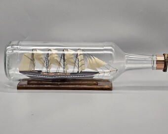 May include: A detailed model of a sailing ship inside a clear glass bottle. The ship has multiple masts with white sails and a dark hull. The bottle rests on a wooden base, and the bottle's neck has a copper-colored stopper.