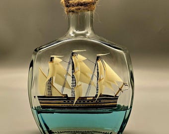Op de afbeelding: Een heldere glazen fles met een houten stop en touwdetails, met een miniatuur zeilschip. Het schip heeft crèmekleurige zeilen en staat in blauwe vloeistof, wat een nautisch thema creëert.