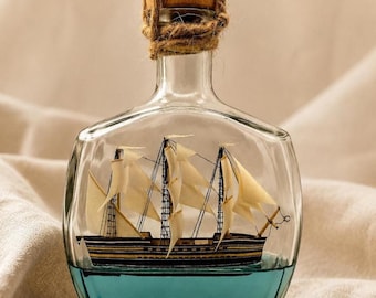 May include: A clear glass bottle containing a miniature model of a sailing ship with white sails. The base of the bottle is filled with a translucent blue liquid. The bottle is sealed with a wooden cork wrapped in natural brown twine.