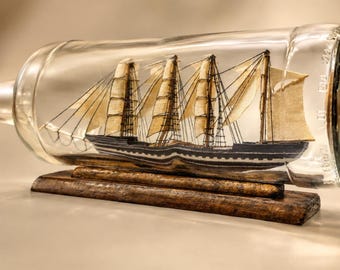May include: A miniature model ship with off-white fabric sails and a dark hull enclosed within a clear glass bottle. The bottle rests horizontally on a rectangular wooden base, showcasing intricate rigging and masts inside the glass vessel.