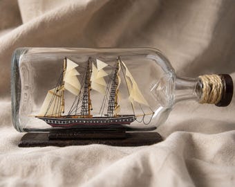 May include: A miniature model ship with cream-colored sails and a dark hull is enclosed within a clear glass bottle. The bottle neck is wrapped in natural jute twine and sealed with a dark brown stopper, resting on a rectangular wooden base.