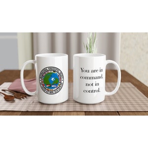May include: Two white ceramic mugs. One mug features a circular logo with the text "International Kriegsspiel Society" and a globe with flags. The other mug has the text "You are in command, not in control."
