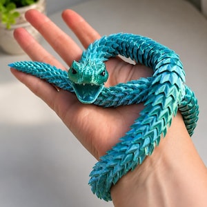 Puede incluir: Una serpiente de juguete flexible y articulada con un patrón de escamas en degradado turquesa y azul. La serpiente tiene una cabeza detallada con la boca abierta, dientes visibles y ojos oscuros, con un diseño de cuerpo segmentado.