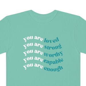 May include: A teal green t-shirt with the text "you are loved, you are strong, you are worthy, you are capable, you are enough" printed in white and teal blue.