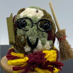 May include: A green, brown, and white yarn creature wearing glasses, a red and yellow scarf, and holding a broom. The creature is sitting on a wooden base.