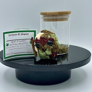 May include: A crocheted plush toy in shades of green, brown, and cream, with large black eyes and a small rolled paper accessory. A clear glass jar with a wooden lid and a certificate of adoption are also present.