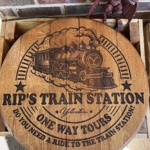 Op de afbeelding: Een ronde houten plaat met de tekst "RIP'S TRAIN STATION" en "ONE WAY TOURS", met een illustratie van een stoomtrein. De tekst bevat de zin "Heeft u een rit naar het treinstation nodig?". De plaat is van lichtbruin hout met een rustieke afwerking.