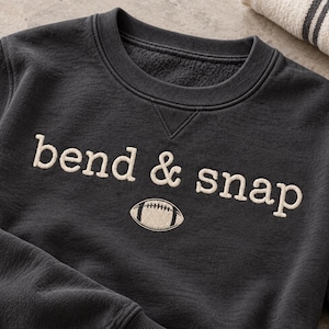 May include: A charcoal gray crewneck sweatshirt featuring the embroidered text "bend & snap" in cream-colored thread. Below the text is an embroidered cream-colored football graphic. The garment has a ribbed collar, cuffs, and hem.