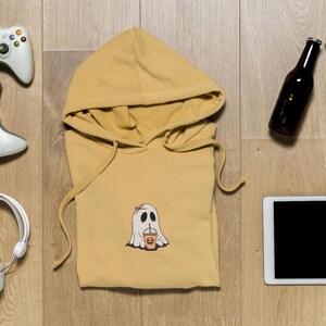 May include: A yellow hoodie with a ghost holding a cup printed on the front. The hoodie is laying flat on a wooden surface with a tablet, two bottles, and two game controllers around it.