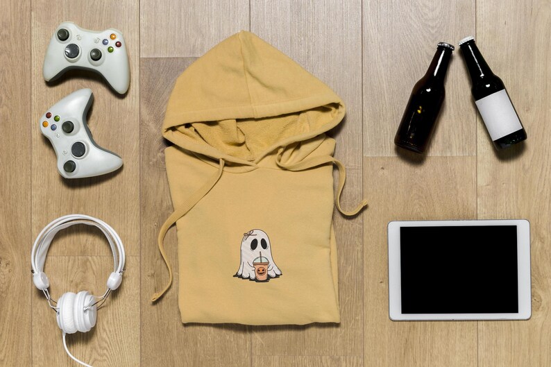 May include: A yellow hoodie with a ghost holding a cup printed on the front. The hoodie is laying flat on a wooden surface with a tablet, two bottles, and two game controllers around it.
