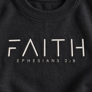 May include: A black crewneck sweatshirt featuring the word FAITH embroidered in white capital letters with a stylised font. Below the word, the text EPHESIANS 2:8 is printed in a smaller, clean white sans-serif font centred on the chest.