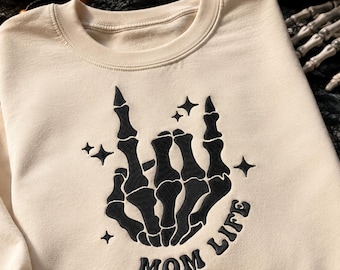 Könnte beinhalten: Ein beigefarbenes Sweatshirt mit Rundhalsausschnitt und einer schwarzen Stickerei einer Skeletthand, die eine Rock-and-Roll-Geste macht. Das Design enthält kleine Sternakzente und den Text "MOM LIFE", der in einer geschwungenen Schrift gestickt ist.
