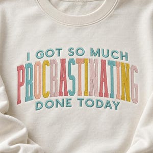 May include: An off-white crewneck sweatshirt featuring embroidered text that reads I GOT SO MUCH PROCRASTINATING DONE TODAY. The word PROCRASTINATING is displayed in large, multi-colored block letters, while the other words are in teal.