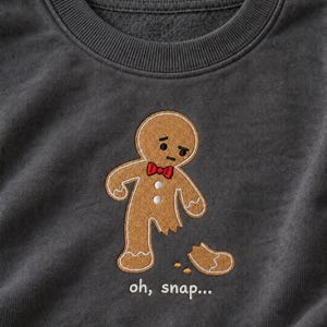 Puede incluir: Una sudadera gris carbón con un bordado de un hombre de jengibre con una pajarita roja y dos botones blancos. Al hombre de jengibre le falta una pierna, con el texto "oh, snap..." bordado en blanco debajo de la figura.