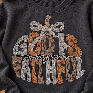 May include: A charcoal gray crewneck sweatshirt featuring an embroidered pumpkin graphic with a bow. The text reads GOD IS FAITHFUL in every season, with letters stitched in alternating white, orange, and brown thread colors.