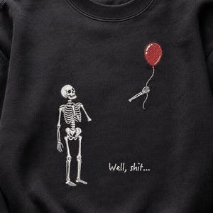 May include: A black crewneck sweatshirt featuring an embroidered design of a white skeleton looking up at a red balloon held by a detached skeletal hand. The text "Well, shit..." is embroidered in white below the balloon and hand.