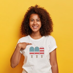 May include: A white t-shirt with a graphic of three ice pops. The ice pops are decorated with red and blue stripes and white sprinkles. The person wearing the shirt is smiling and pointing at the graphic.