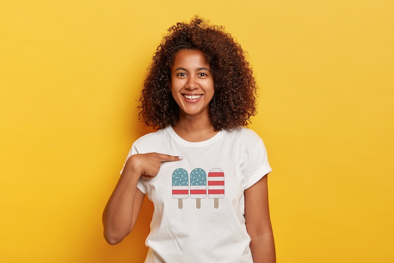 May include: A white t-shirt with a graphic of three ice pops. The ice pops are decorated with red and blue stripes and white sprinkles. The person wearing the shirt is smiling and pointing at the graphic.