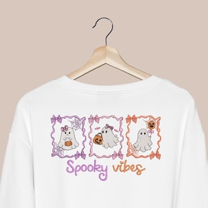 K&ouml;nnte beinhalten: Ein wei&szlig;es Langarmshirt mit einem Grafikdruck von drei rechteckigen Rahmen, die Cartoon-Geister mit Schleifen, K&uuml;rbissen und Spinnenweben enthalten. Unter den Rahmen ist der Text Spooky vibes in einer lila und orangefarbenen Schrift gedruckt.