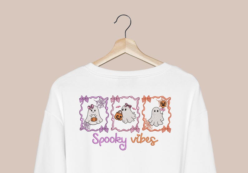 K&ouml;nnte beinhalten: Ein wei&szlig;es Langarmshirt mit einem Grafikdruck von drei rechteckigen Rahmen, die Cartoon-Geister mit Schleifen, K&uuml;rbissen und Spinnenweben enthalten. Unter den Rahmen ist der Text Spooky vibes in einer lila und orangefarbenen Schrift gedruckt.