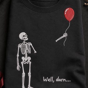 May include: A black crewneck sweatshirt featuring white embroidery of a skeleton standing and looking up at a detached skeletal hand holding a red balloon. The text "Well, darn..." is embroidered in white below the skeleton on the front.