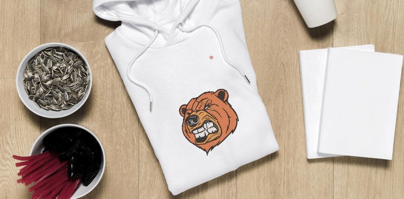 May include: White hooded sweatshirt with a brown bear face graphic. The bear has a fierce expression with sharp teeth and a furrowed brow.