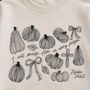 Pode incluir: Uma camisola de gola redonda bege com bordado de linhas pretas de várias abóboras, dois laços, duas folhas e uma cruz. O texto diz: I will praise Him in every season Psalm 146:2. O design está centrado na parte da frente da peça.