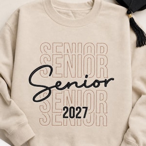 May include: A beige crewneck sweatshirt featuring the word SENIOR embroidered in a repeating stacked pattern with brown thread. The word Senior is also embroidered in a bold black script font, and the number 2027 is embroidered in black.