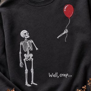 May include: A black crewneck sweatshirt featuring white embroidery of a skeleton looking up at a detached skeletal hand holding a red embroidered balloon. The text "Well, crap..." is embroidered in white below the hand on the front of the garment.