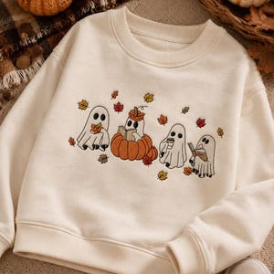 May include: A cream-colored crewneck sweatshirt featuring an embroidered graphic of four white ghosts. One ghost sits inside a large orange pumpkin, while the others hold books, a coffee cup, and autumn leaves, surrounded by scattered fall foliage.