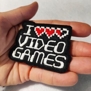May include: A black embroidered patch featuring the white text I, three red pixelated heart icons, and the white text VIDEO GAMES. The patch has a rectangular shape with rounded corners and a thick black border, showcasing detailed stitching.