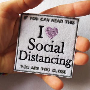Pode incluir: Patch quadrado branco com texto bordado em preto: "Se consegues ler isto, adoro distanciamento social. Estás muito perto". Um coração roxo é bordado acima de "distanciamento social". O patch tem uma borda cinzenta.