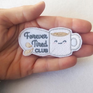 Puede incluir: Un parche bordado con una taza de café blanca con una cara sonriente y una burbuja de diálogo que dice "Forever Tired Club". El parche tiene un borde gris y detalles dorados.