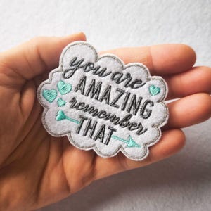 Puede incluir: Parche de fieltro blanco en forma de nube con las palabras "you are AMAZING remember THAT" bordadas en negro y verde azulado. El parche tiene un borde plateado y corazones y un diseño de flecha verde azulado.