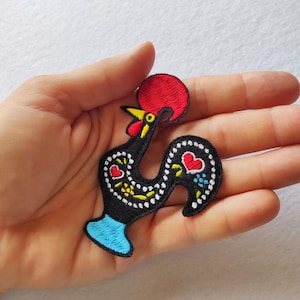 Puede incluir: Parche bordado del Gallo de Barcelos, un símbolo de Portugal. El gallo negro tiene una cresta roja, un pico amarillo y patas azules. El cuerpo del gallo está decorado con corazones y motivos florales.