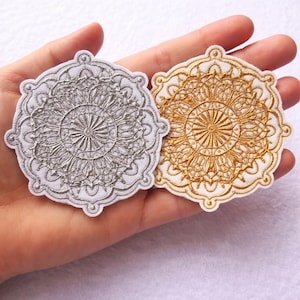 Pode incluir: Dois patches de mandala bordados. Um é prateado, o outro dourado, ambos com designs intrincados. Os patches são circulares com uma borda branca e provavelmente são para fins decorativos.