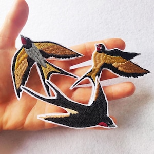 Pode incluir: Patches bordados com andorinhas em voo. Os patches apresentam costuras detalhadas em preto, castanho, dourado e vermelho. As andorinhas têm contornos brancos, criando um contraste marcante. Ideais para adicionar um toque decorativo a roupas ou acessórios.