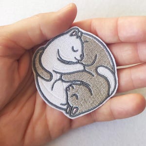Könnte beinhalten: Gestickter Aufnäher mit einem Yin-Yang-Design aus zwei Katzen. Eine Katze ist weiß, die andere ist hellbraun, beide mit schwarzen Umrissen. Der Aufnäher ist kreisförmig und hat einen weißen Rand.