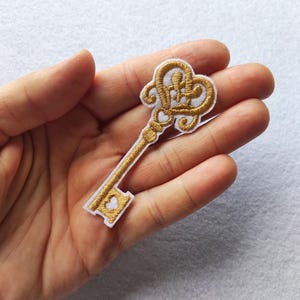 Puede incluir: Parche de llave dorada bordada con reverso blanco. La llave presenta detalles ornamentados, incluido un ojo de cerradura en forma de corazón y remolinos decorativos. Este parche es adecuado para agregar a ropa, bolsos u otros artículos de tela.