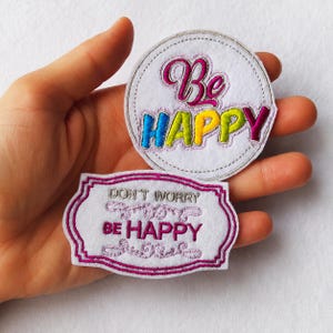 Könnte beinhalten: Zwei weiße Stoffaufnäher mit gesticktem Text. Der runde Aufnäher zeigt die Worte Be HAPPY in Pink, Blau, Gelb und Grün. Der rechteckige Aufnäher mit gewelltem Rand zeigt den Text DON'T WORRY BE HAPPY in Pink und Grau.