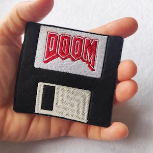 May include: A square embroidered patch shaped like a black floppy disk. The top section features a white label with the word DOOM stitched in bold red letters. The bottom section includes a white rectangular area representing the disk's sliding metal shutter.
