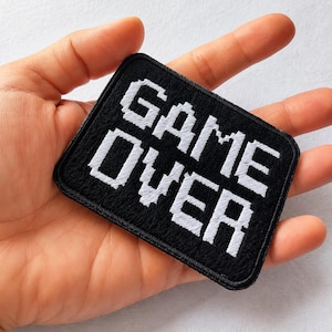 May include: A rectangular embroidered patch with a black background and white pixelated text that reads GAME OVER. The patch features a stitched border and a textured fabric surface, designed with a classic retro video game aesthetic.