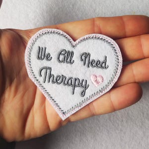 Könnte beinhalten: Ein weißes herzförmiges Patch mit dem gestickten Text "We All Need Therapy" in schwarzer Schrift. Das Patch hat einen rosa Rand und ein kleines rosa Herz. Das Patch wird in einer Hand gehalten.