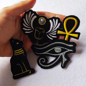 May include: Embroidered patches featuring Egyptian symbols: a black cat with yellow accents, a scarab beetle, an ankh, and the Eye of Horus. The patches are black with white and yellow details, suitable for clothing or accessories.